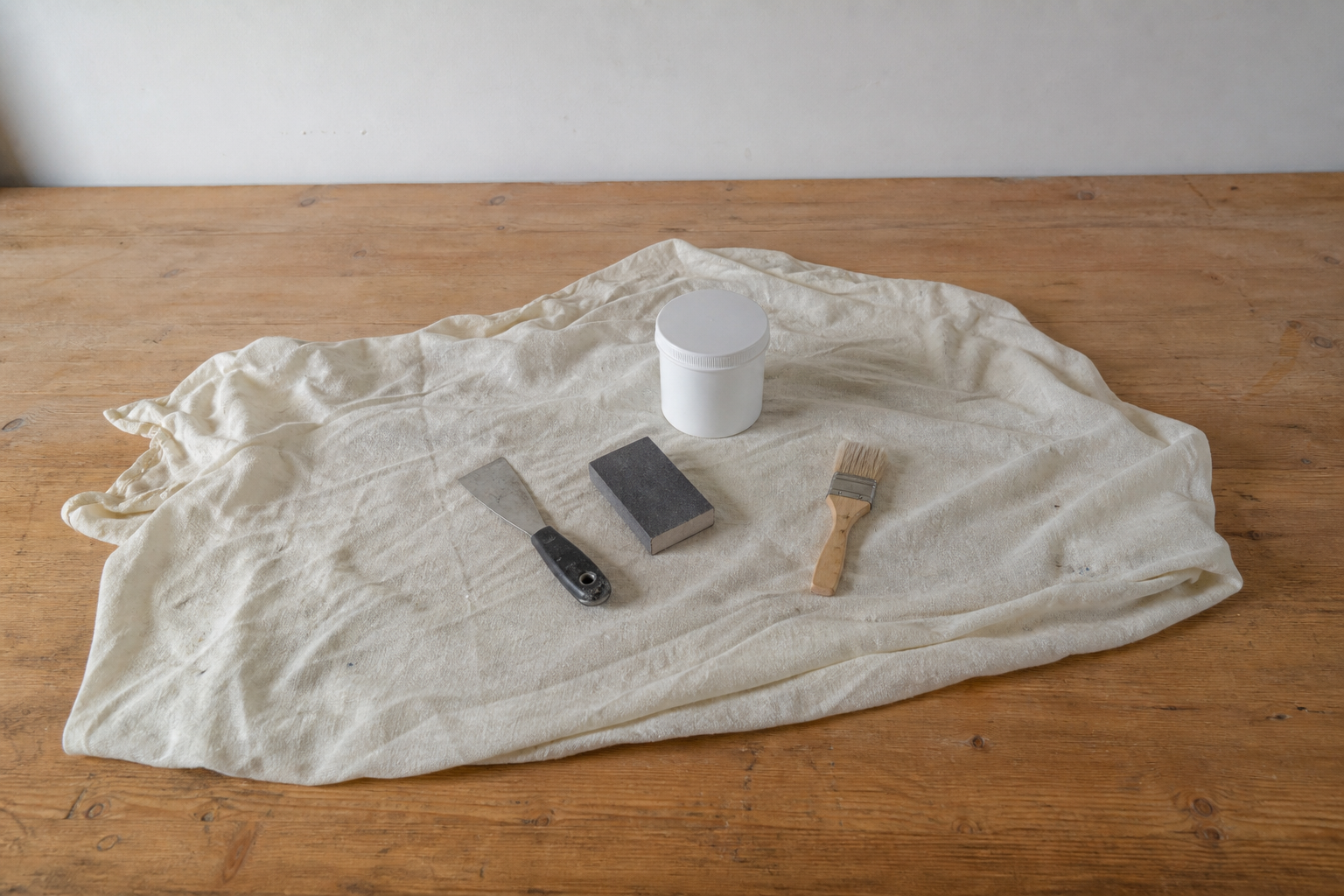 Asymmetric patch-preparation tools—a putty knife, sanding block, brush, and blank repair container—resting on a rumpled drop cloth on wood.